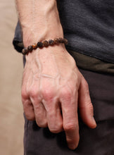 Load image into Gallery viewer, A close-up of a person's hand and wrist wearing a beaded bracelet made of brown and dark stone beads, paired with a dark gray shirt and dark pants.
