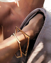 Load image into Gallery viewer, A close-up of a person's wrist wearing two overlapping geometric gold bangles, with a textured gray garment draped around the arm in warm sunlight.
