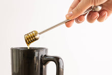 Load image into Gallery viewer, Honey being drizzled from a honey dipper into a black mug on a light background
