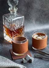 Load image into Gallery viewer, Two glasses with leather sleeves filled with a drink, next to a decanter on a dark surface.
