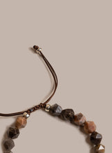 Load image into Gallery viewer, A close-up of a beaded cord necklace featuring faceted brown, beige, and dark gray stones, accented with small metallic silver beads, on a solid beige background.
