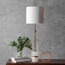 Load image into Gallery viewer, Table lamp with white shade and gold accents on a wooden surface against a gray wall.
