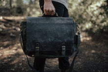 Load image into Gallery viewer, A person holding a black leather messenger bag with a zippered front pocket and an adjustable shoulder strap.
